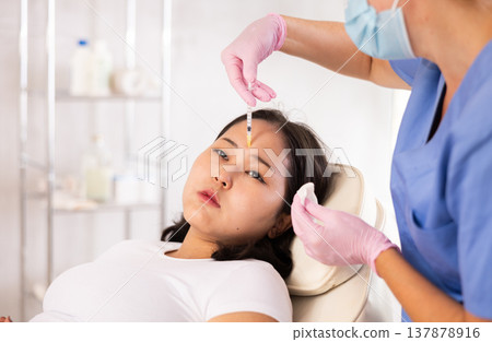 Face of young female client receiving injections during lip enhancement procedure, professional cosmetologist hands in rubber gloves holding syringe Face of young female client receiving injections during lip enhancement procedure, professional cosmetologist hands in rubber gloves holding syringe 137878916