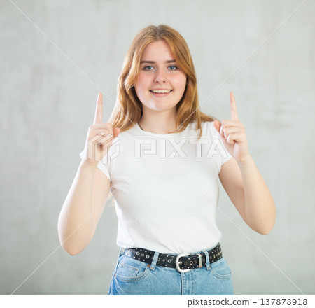 Portrait of cheerful charismatic teen girl in white t-shirt and blue jeans pointing up against gray wall with copy space Portrait of cheerful charismatic teen girl in white t-shirt and blue jeans pointing up against gray wall with copy space 137878918