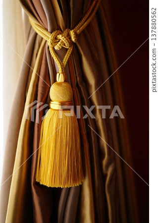Golden curtain tassel tied with braided cord against rich draped fabric in warm light 137880562