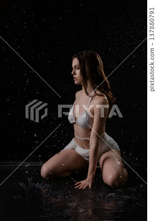 Sensual woman in a white swimsuit kneeling in falling water on a dark, reflective surface. Sensual woman in a white swimsuit kneeling in falling water on a dark, reflective surface. 137880791