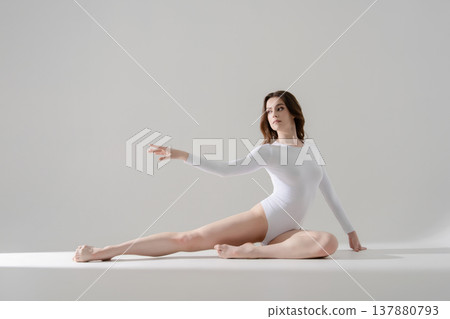 Elegant woman in white leotard posing gracefully on floor in a bright minimalist studio. Elegant woman in white leotard posing gracefully on floor in a bright minimalist studio. 137880793