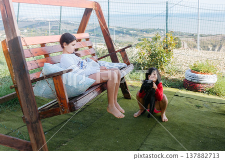 a girl sitting on a garden swing on a house patio another girl sitting next to her with a cat in hands a girl sitting on a garden swing on a house patio another girl sitting next to her with a cat in hands 137882713