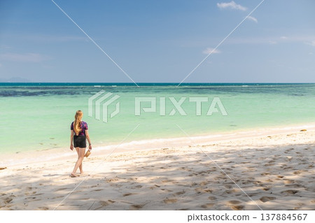 Young woman walking barefoot along sandy beach in Koh Samui, Thailand, holding hat and looking toward turquoise sea under clear blue sky during tropical summer travel Young woman walking barefoot along sandy beach in Koh Samui, Thailand, holding hat and looking toward turquoise sea under clear blue sky during tropical summer travel 137884567