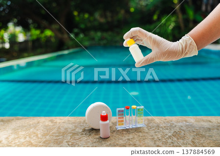 Gloved hand holding reagent bottle above swimming pool with test kit and chlorine tablet on pool edge, water quality control and pool maintenance concept with tropical background Gloved hand holding reagent bottle above swimming pool with test kit and chlorine tablet on pool edge, water quality control and pool maintenance concept with tropical background 137884569