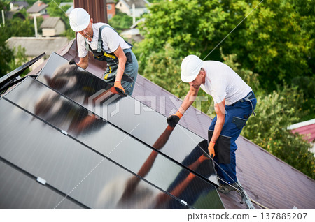 Workers building solar panel system on rooftop of house. Two men installers in helmets installing photovoltaic solar module outdoors. Alternative, green and renewable energy generation concept. Workers building solar panel system on rooftop of house. Two men installers in helmets installing photovoltaic solar module outdoors. Alternative, green and renewable energy generation concept. 137885207