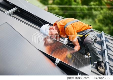 Worker building solar panel system on rooftop of house. Man engineer in helmet installing photovoltaic solar module outdoors. Alternative, green and renewable energy generation concept. Worker building solar panel system on rooftop of house. Man engineer in helmet installing photovoltaic solar module outdoors. Alternative, green and renewable energy generation concept. 137885209