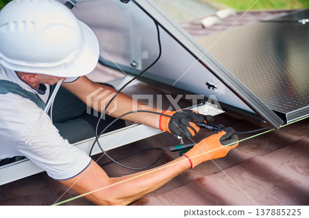 Worker installing solar panels system on rooftop of house. Electrician wiring solar modules together for generating electricity through photovoltaic effect. Concept of alternative, renewable energy. Worker installing solar panels system on rooftop of house. Electrician wiring solar modules together for generating electricity through photovoltaic effect. Concept of alternative, renewable energy. 137885225