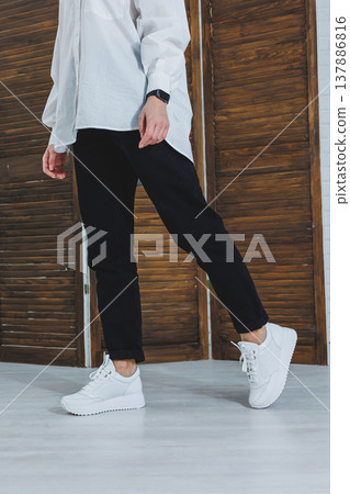 Close-up of female legs in black jeans in stylish leather white sneakers. Fashionable woman in new sneakers. Modern seasonal collection of stylish sneakers. Women's fashion. 137886816