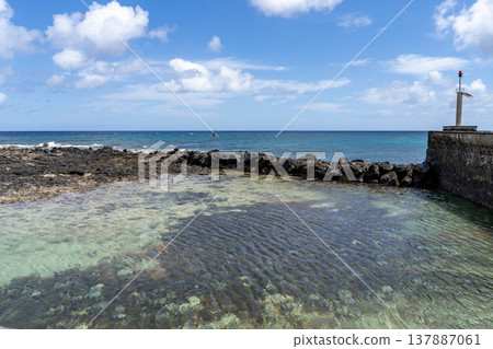 View of a natural pool in Lanzarote, with the sea and sky in the background. A lighthouse is present on the right side. View of a natural pool in Lanzarote, with the sea and sky in the background. A lighthouse is present on the right side. 137887061