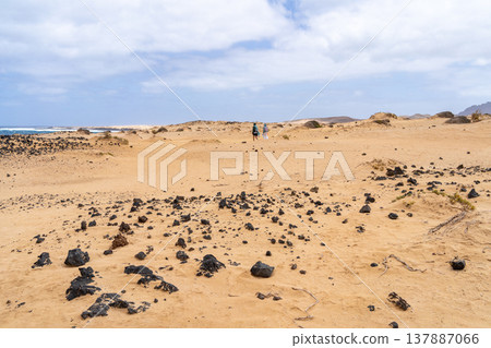 Two people walking on a sandy landscape with black rocks scattered around. La Graciosa, Lanzarote. The sky is cloudy. Two people walking on a sandy landscape with black rocks scattered around. La Graciosa, Lanzarote. The sky is cloudy. 137887066