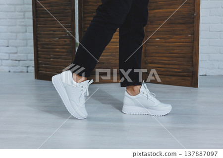 Beautiful women's shoes with legs, leather white sneakers, in the interior of a room or office, shoes for a woman, black trousers and white sneakers, casual and modern style, modern and trendy Beautiful women's shoes with legs, leather white sneakers, in the interior of a room or office, shoes for a woman, black trousers and white sneakers, casual and modern style, modern and trendy 137887097