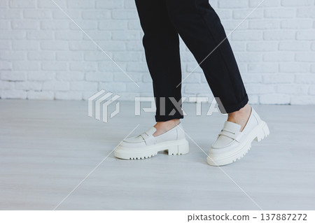 Legs of a young beautiful woman in black trousers and modern stylish white leather loafers. New collection of women's shoes spring-summer 2022 Legs of a young beautiful woman in black trousers and modern stylish white leather loafers. New collection of women's shoes spring-summer 2022 137887272