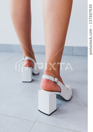 Smooth beautiful slender legs of a girl in white leather high-heeled sandals on the floor against a gray parquet floor. flawless hair removal and tanning, female beauty 137887630