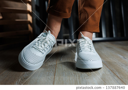 Close-up of female legs in white perforated leather sneakers. Women's summer sneakers. Collection of women's leather shoes Close-up of female legs in white perforated leather sneakers. Women's summer sneakers. Collection of women's leather shoes 137887640