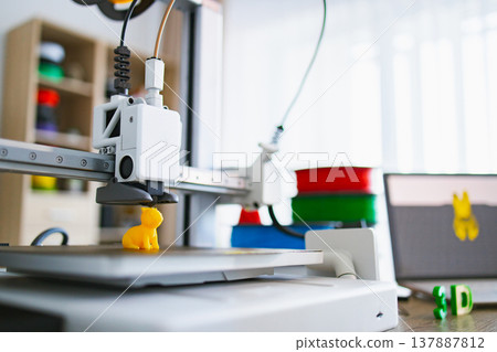 A modern 3D printer is actively creating a small yellow figurine on its build plate, with spools of filament and a laptop in the background 137887812