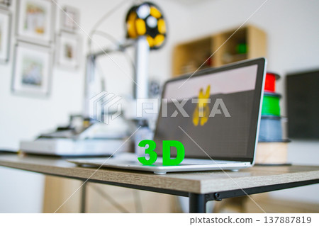 A laptop displays a 3D model next to a 3D printer and spools of filament, showcasing a modern creation space 137887819