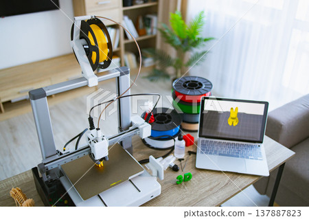 A 3D printer is set up on a desk with multiple spools of filament and a laptop displaying a 3D model A 3D printer is set up on a desk with multiple spools of filament and a laptop displaying a 3D model 137887823