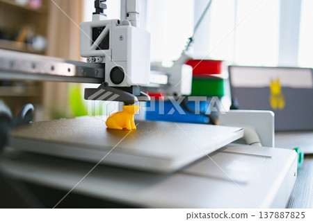 A modern 3D printer is actively creating a small yellow figurine on its build plate with filament spools in the background 137887825
