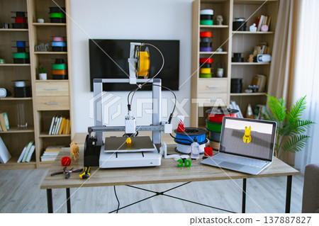 A 3D printer is actively creating a yellow object on a desk with spools of filament, tools, and a laptop displaying a 3D model A 3D printer is actively creating a yellow object on a desk with spools of filament, tools, and a laptop displaying a 3D model 137887827