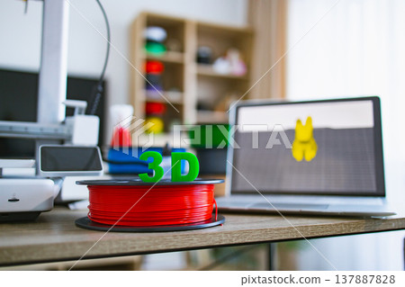 A 3D printer, filament spool, and laptop with a 3D model displayed are arranged on a wooden desk, ready for creation 137887828