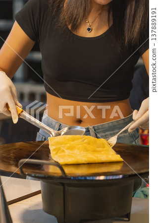 A woman prepares sweet pancake roti at a street stall in Bangkok. She uses two spoons to shape the dough on a hot pan. This scene shows local food culture and cooking methods in Thailand. High quality A woman prepares sweet pancake roti at a street stall in Bangkok. She uses two spoons to shape the dough on a hot pan. This scene shows local food culture and cooking methods in Thailand. High quality 137889191