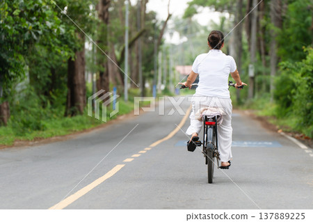 a woman riding a bicycle on an asphalt road, rear view, which passes through a wooded or park area in summer. Copy space on the left, she is dressed in light clothes. Freedom and relaxation - A quiet 137889225