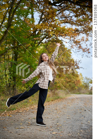 Happy young woman posing playfully on rural road under autumn trees, full body lifestyle portrait with colorful foliage, concept of freedom, joy and carefree mood Happy young woman posing playfully on rural road under autumn trees, full body lifestyle portrait with colorful foliage, concept of freedom, joy and carefree mood 137889368