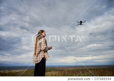 Woman piloting drone in cloudy sky over open landscape. Side view of female operator with controller on hill, aerial photography and exploration concept of technology 137889394