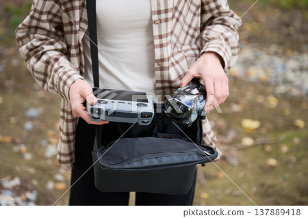Woman holds drone and controller taken from bag. Close up of hands preparing quadcopter outdoors, organizing gear before flight, concept of hobby, readiness and technology Woman holds drone and controller taken from bag. Close up of hands preparing quadcopter outdoors, organizing gear before flight, concept of hobby, readiness and technology 137889418