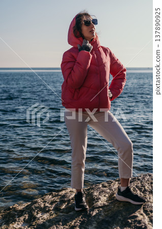 Woman puffer jacket sea standing on a rocky shore wearing a pink coat and sunglasses enjoying the winter sun Woman puffer jacket sea standing on a rocky shore wearing a pink coat and sunglasses enjoying the winter sun 137890925
