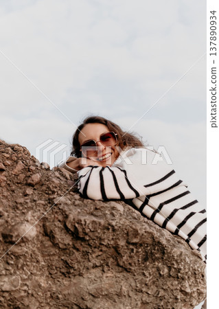 Woman sweater outdoors resting on a brown rock against a bright sky while smiling contentedly and wearing sunglasses Woman sweater outdoors resting on a brown rock against a bright sky while smiling contentedly and wearing sunglasses 137890934