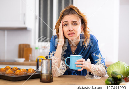 Young woman with headache standing with cup of coffee in home kitchen Young woman with headache standing with cup of coffee in home kitchen 137891993