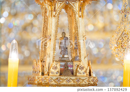 Golden shrine with small buddha statue placed at center. Decorative structure and candle light create warm religious setting. Golden shrine with small buddha statue placed at center. Decorative structure and candle light create warm religious setting. 137892429