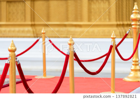 Golden stanchions with red velvet rope arranged along a red carpet pathway creating a controlled access setup for luxury events and crowd management. Golden stanchions with red velvet rope arranged along a red carpet pathway creating a controlled access setup for luxury events and crowd management. 137892821