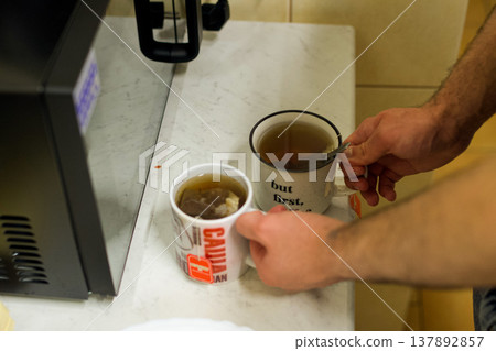 A man pours tea into two mugs on the kitchen table. The concept of comfort, hospitality, and everyday life, morning rituals, warmth, relaxation, and a homey lifestyle. A man pours tea into two mugs on the kitchen table. The concept of comfort, hospitality, and everyday life, morning rituals, warmth, relaxation, and a homey lifestyle. 137892857