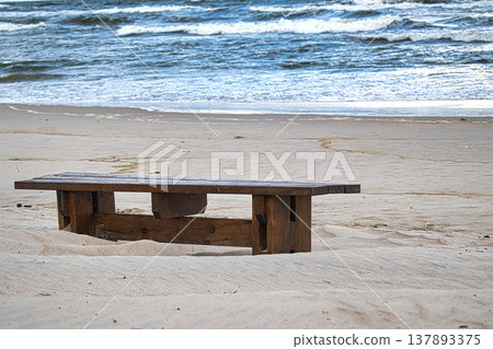 Weathered wooden bench sits alone on a sandy beach overlooking the ocean waves Weathered wooden bench sits alone on a sandy beach overlooking the ocean waves 137893375
