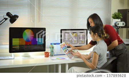Two designers collaborate on mobile app UX UI, reviewing color palettes and wireframes on desktop screens in a modern creative office Two designers collaborate on mobile app UX UI, reviewing color palettes and wireframes on desktop screens in a modern creative office 137893628