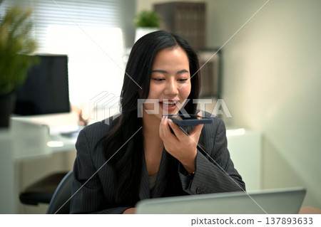 Professional woman using voice assistant chatbot voice on smartphone while working on laptop in modern office Professional woman using voice assistant chatbot voice on smartphone while working on laptop in modern office 137893633