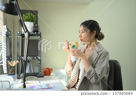 Businesswoman eating instant noodles at office desk, casual lunch break during workday in modern workspace. 137893664