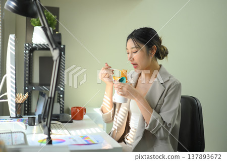 Businesswoman eating instant noodles at office desk, casual lunch break during workday in modern workspace. 137893672