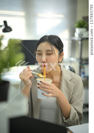 Businesswoman eating instant noodles at office desk, casual lunch break during workday in modern workspace. Businesswoman eating instant noodles at office desk, casual lunch break during workday in modern workspace. 137893795