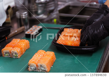 Chef Packing Salmon Sushi Into Black Container On Prep Table, Gloved Hand Placing Rolls 137894630