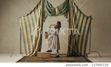 Young girl playing saxophone standing on box in studio with theatrical drapery. Young girl playing saxophone standing on box in studio with theatrical drapery. 137895130