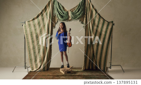 Smiling young girl holding violin on small stage platform with curtains. Smiling young girl holding violin on small stage platform with curtains. 137895131