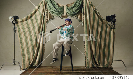 Young boy playing clarinet sitting on stool in studio stage with vintage curtains. Young boy playing clarinet sitting on stool in studio stage with vintage curtains. 137895142