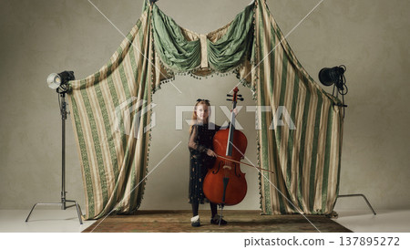 Young girl holding cello on studio stage with curtains and lighting equipment behind scenes setup. Young girl holding cello on studio stage with curtains and lighting equipment behind scenes setup. 137895272