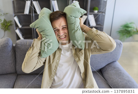 Annoyed man with ear sensitivity holding pillows to block sound of noisy neighbors Annoyed man with ear sensitivity holding pillows to block sound of noisy neighbors 137895831