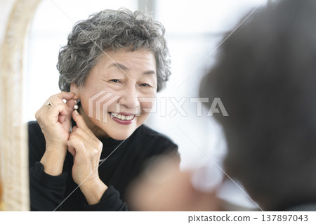 A smiling senior woman puts on earrings while looking in the mirror. 137897043