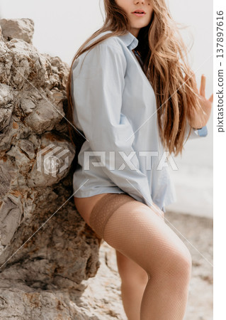 Woman stockings beach posing against rocks wearing blue oversized shirt outdoors near ocean during daylight 137897616
