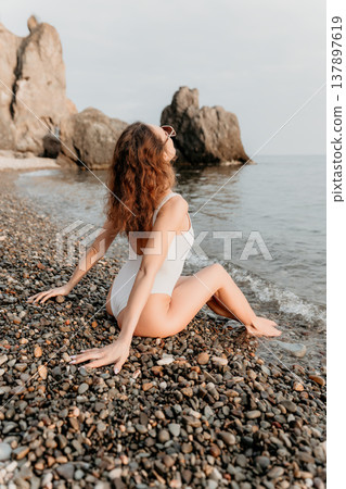 Woman beach swimsuit sitting on a pebble shore looking at the sea and rocky cliffs during summer vacation 137897619